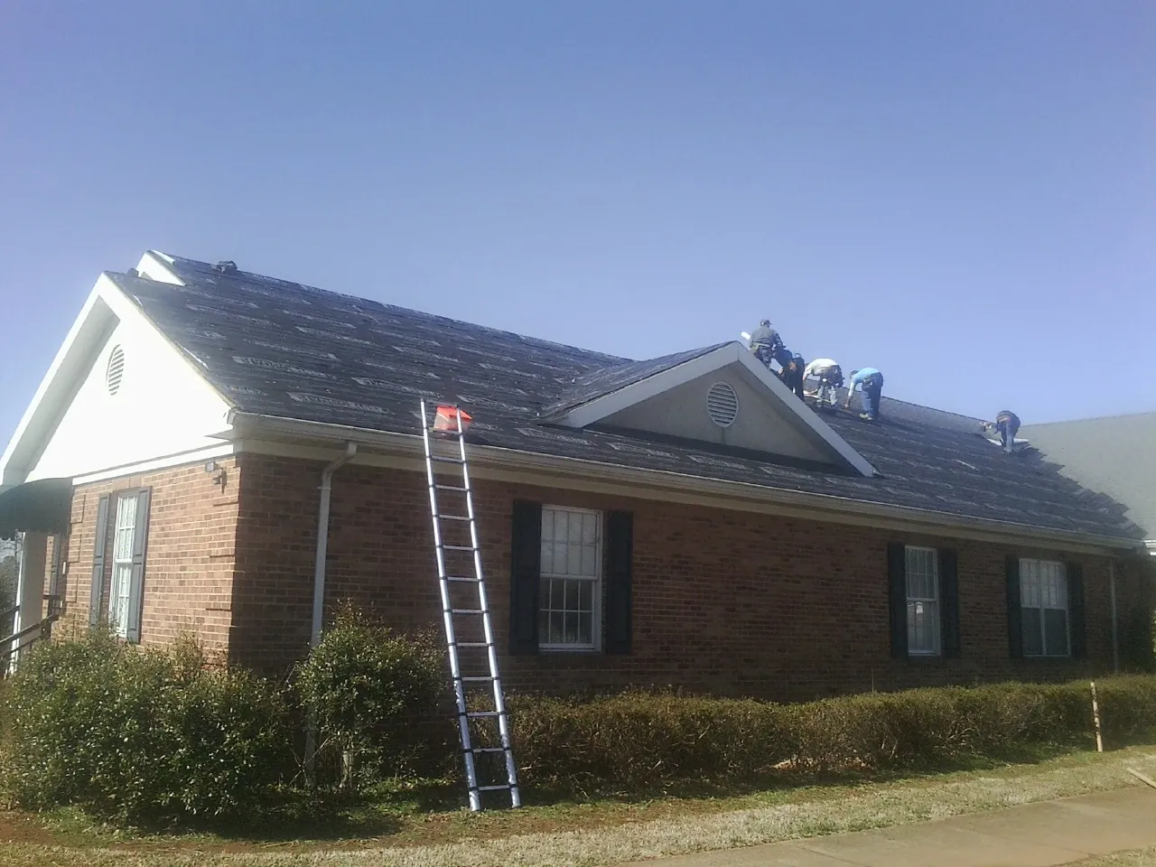 Crew tearing shingles off a brick ranch roof with a ladder set — Emergency Roof Repair work by our crew in Prince Frederick