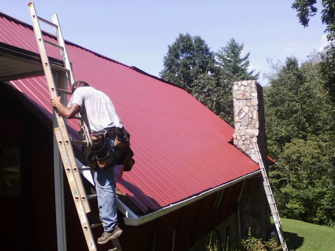 Roofer on a ladder beside a red metal roof and stone chimney — Soffit & Fascia Repair by Roofing Brothers in Prince Frederick